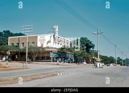 Vue sur la rue Tom Lawrence, White River (Witrivier), province de Mpumalanga, Afrique du Sud en 1962. La ville est une petite ville de vacances et d'agriculture située juste au nord de Mbombela dans l'ancienne province de Transvaal. Les fermes de la région produisent des fruits tropicaux, des légumes, des fleurs et du bois. Le bâtiment situé à l'angle de la rue William Lynn indique que le Kruger Park café y était situé. Un grand panneau d'affichage Coca Cola présente une girafe. Cette image provient d'une transparence de couleur 35 mm amateur ancienne. Banque D'Images