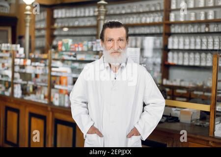 Portrait d'un beau pharmacien de sexe masculin âgé, portant un manteau blanc, debout devant les étagères de médicaments de l'ancienne pharmacie. Banque D'Images