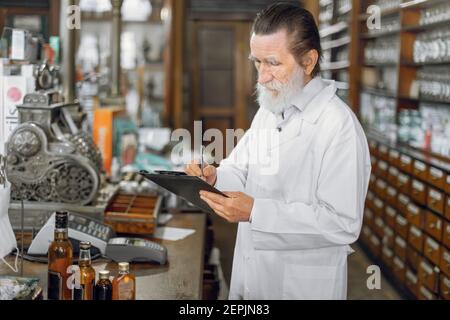 Portrait d'un pharmacien senior expérimenté concentré écrit sur un planchette à pince tout en se tenant dans un bel intérieur ancien de pharmacie Banque D'Images