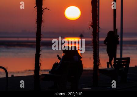 Southend on Sea, Essex, Royaume-Uni. 27 février 2021. La chaude journée ensoleillée s'est terminée par un coucher de soleil lumineux derrière les palmiers et les visiteurs le long du front de mer à Southend on Sea. Les gens marchent et s'assoyent sur la promenade de l'Esplanade de l'Ouest pendant que le soleil se couche pendant le confinement de la COVID 19. Deux femelles assises et mangeant de la crème glacée Banque D'Images