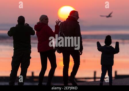 Southend on Sea, Essex, Royaume-Uni. 27 février 2021. La chaude journée ensoleillée s'est terminée par un coucher de soleil lumineux derrière les palmiers et les visiteurs le long du front de mer à Southend on Sea. Les gens marchent et s'assoyent sur la promenade de l'Esplanade de l'Ouest pendant que le soleil se couche pendant le confinement de la COVID 19. La famille est à l'extérieur pour une promenade le soir Banque D'Images