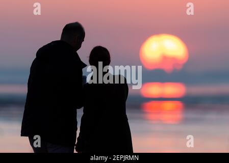 Southend on Sea, Essex, Royaume-Uni. 27 février 2021. La chaude journée ensoleillée s'est terminée par un coucher de soleil lumineux derrière les palmiers et les visiteurs le long du front de mer à Southend on Sea. Les gens marchent et s'assoyent sur la promenade de l'Esplanade de l'Ouest pendant que le soleil se couche pendant le confinement de la COVID 19. Couple mâle et femelle regardant le soleil descendre Banque D'Images