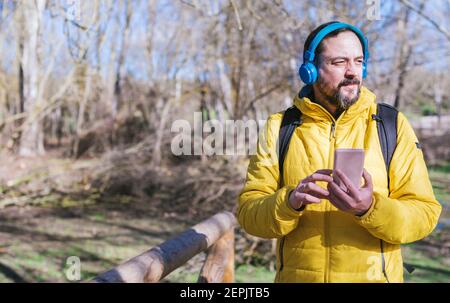 un homme de taille basse avec une barbe qui écoute de la musique avec un casque et un téléphone portable tout en marchant par temps ensoleillé. Banque D'Images