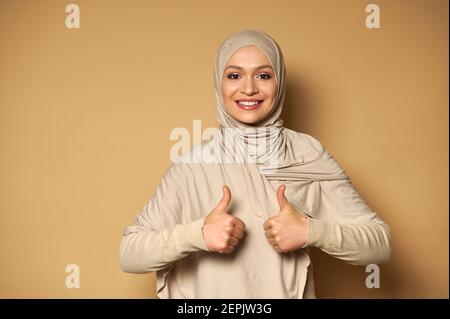 Une femme musulmane mignonne avec une tête couverte dans le hijab montre des pouces sourit avec un sourire éclatant sur fond beige avec copier l'espace Banque D'Images