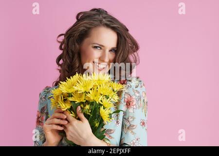 femme moderne souriante avec de longs cheveux ondulés brunette avec des fleurs de chrysanthèmes jaunes isolées sur rose. Banque D'Images