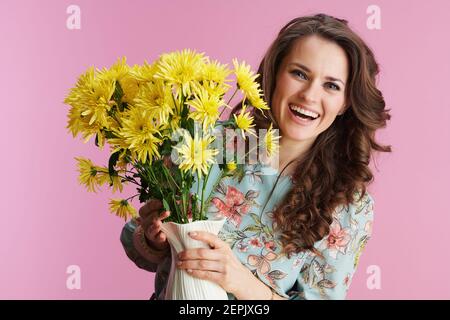 Portrait d'une femme heureuse d'âge moyen avec de longs cheveux de brunette ondulés avec des fleurs de chrysanthèmes jaunes dans un vase isolé sur fond rose. Banque D'Images