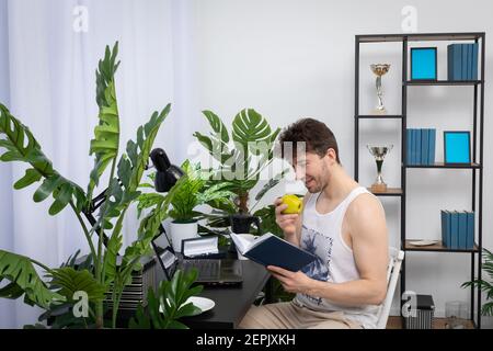 Un employé s'assoit à la maison et examine un calendrier de livre avant de commencer à travailler sur son ordinateur portable. Travail à distance pendant une pandémie. Un salon lumineux dans un style moderne avec un espace préférable. Banque D'Images