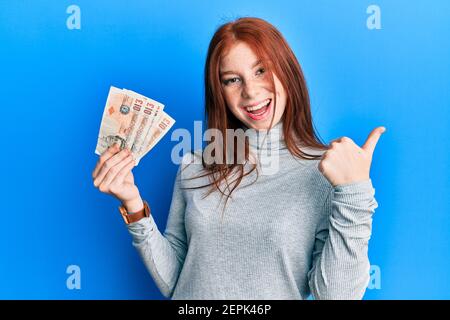 Jeune fille à tête rouge détenant 10 billets de banque au royaume-uni en pointant le pouce vers le haut, souriant et ouvert bouche Banque D'Images