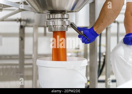 Gros plan sur la main d'un inconnu portant un gant de protection ouverture des silos avec de la crème ou de la marmelade lors du versement en usine dans l'ensemble seau - nourriture Banque D'Images