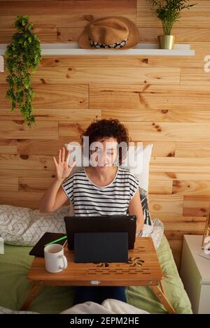 Une femme aux cheveux bouclés lève la main pour saluer ses collègues lors d'une réunion d'affaires. Elle est assise sur son lit avec son dos à un beau mur en bois. Banque D'Images