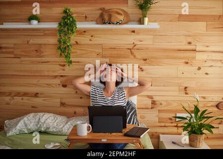 Une jeune femme aux cheveux bouclés étire le dos pour la dégager de l'esprit. Elle est assise sur son lit et travaille de la maison. Dans la chambre il y a un mur en bois. S Banque D'Images