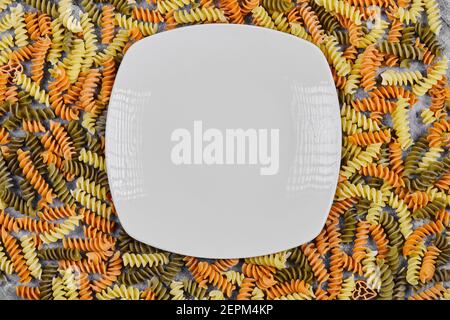 Pâtes fusilli crues colorées éparpillées sur une table en marbre avec plaque blanche Banque D'Images