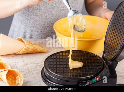 Une femme utilise un gaufrier électrique anti-adhésif pour fabriquer des cornets de glace faits maison. Des cônes fraîchement roulés à la main sont posés sur un comptoir en marbre Banque D'Images