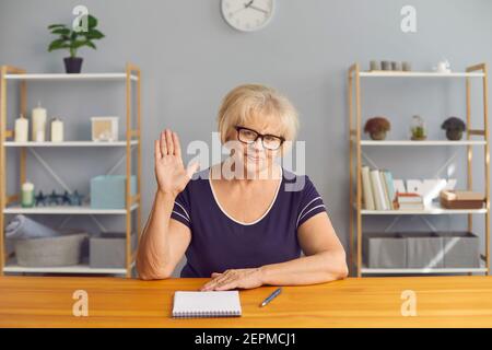 Une femme sénior, blogueuse et couronnée de succès, fait des vagues de sa main tout en étant assise devant l'appareil photo à une table. Banque D'Images