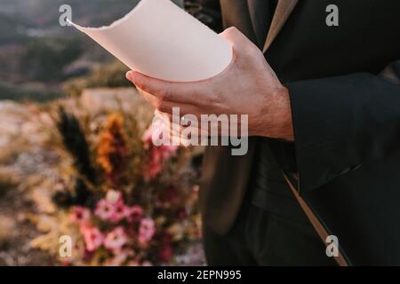 Fiancé non reconnaissable portant un costume noir debout avec du papier pendant le mariage voeu dans la nature contre les bouquets colorés de fleurs sur flou arrière-plan Banque D'Images
