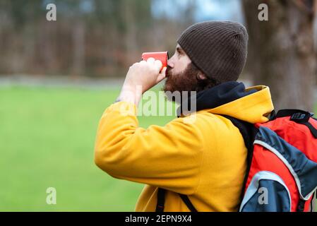 Vue latérale d'un trekker mâle barbu avec un sac à dos pour boire du thé de la tasse en métal sur la prairie Banque D'Images