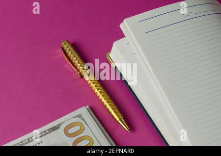 Journal ouvert, stylo doré et pile de cent dollars américains sur fond rose. Vue de dessus inclinée. Mise au point sélective Banque D'Images