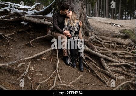 Corps complet de couple doux positif assis près de l'arbre et embrasser et s'embrasser pendant une journée romantique dans les bois Banque D'Images