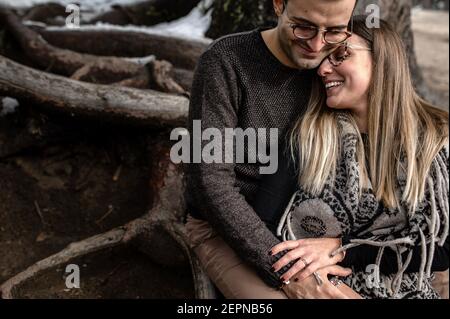 Corps complet de couple doux positif assis près de l'arbre et se embrasser pendant une journée romantique dans les bois Banque D'Images