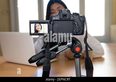 Rognez une femme ethnique souriante avec l'enregistrement de vidéos sur netbook sur professionnel caméra à table dans la maison Banque D'Images