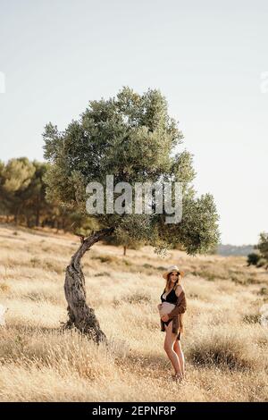 Femme enceinte élégante debout près d'un arbre placé dans le champ avec de l'herbe sèche et touchant le ventre par beau temps Banque D'Images