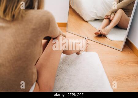 Vue arrière de la jeune femme pensive touchant la jambe contre le miroir assis sur le parquet de la maison Banque D'Images