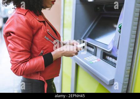 Anonyme afro-américaine femme debout près de ATM et insertion de crédit tout en souriant et en regardant loin Banque D'Images