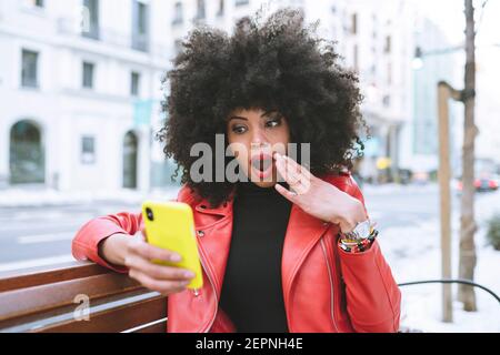 Élégante femme afro-américaine étonnante lisant des informations sur le téléphone portable avec bouche ouverte assise sur banc en ville Banque D'Images