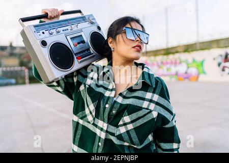 Jeune femme tendance en lunettes de soleil reflétant ciel nuageux à l'écoute chanson de l'ancien magnétophone contre le mur de graffiti en plein soleil Banque D'Images