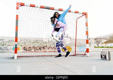 Femme gaie montrant un geste de corne tout en faisant du pigeyback sur un ami afro-américain sur un terrain de sport avec un magnétophone rétro Banque D'Images