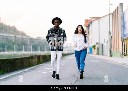 Les jeunes sont des partenaires multiraciaux dans des vêtements et des lunettes de soleil à la mode qui parlent tout en regardant la caméra sur la route de la ville Banque D'Images