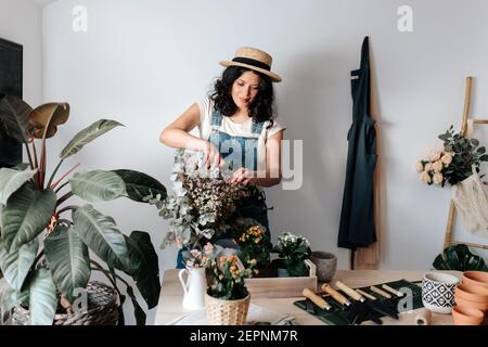 Jeune horticulteur femelle en chapeau de paille coupant le feuillage de la plante près fleurs en pot sur la table avec divers outils à la maison Banque D'Images