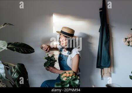 Jeune femme jardinier en tenue décontractée en utilisant l'instrument de jardinage sur fleurs fleuries de plantes en pot à la maison Banque D'Images