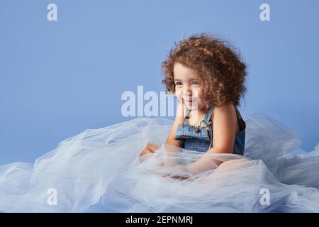 Adorable fille à poil dur souriant mignon assis sur un tissu fin sur fond bleu et en regardant loin Banque D'Images