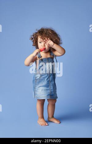 Tout le corps de la petite fille bouleversée sur les vêtements d'été barefooted debout avec de la crème glacée sur fond bleu studio Banque D'Images