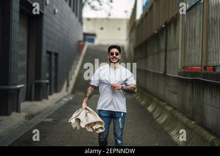 Jeune heureux tatoué ethnique mâle dans des lunettes de soleil et des jeans déchirés promenade sur la passerelle entre les bâtiments urbains Banque D'Images