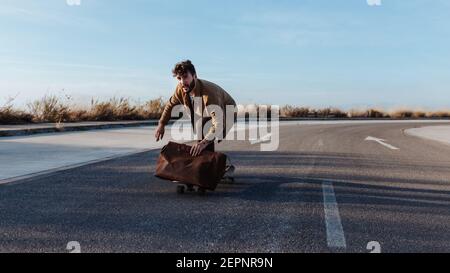 Patineuse masculine pleine corps sérieuse jeune barbu dans des vêtements tendance avec sac en cuir, planche à roulettes le long de la route asphaltée Banque D'Images