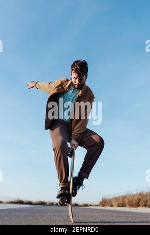 Patineuse masculine à corps entier, jeune et barbu, debout sur le bord de planche à roulettes pour garder l'équilibre tout en effectuant des tours sur route en asphalte avec lever et regarder à la main Banque D'Images