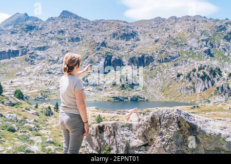 Vue arrière femelle anonyme Backpacker prenant des photos sur le smartphone de Superbes montagnes pierreuses avec lac dans la vallée de Ruda en catalan Pyrénées Banque D'Images