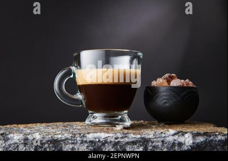 Verre transparent de café noir fort avec mousse près du bol de cubes de sucre brun sur fond noir Banque D'Images