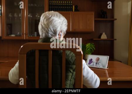Vue arrière de la patiente âgée anonyme dans les oreillettes PSID parler avec un médecin sur une tablette pendant un appel vidéo à la maison Banque D'Images