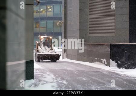 Une petite pelle hydraulique avec un godet élimine la neige d'une route dans une rue urbaine lors d'une forte chute de neige. Banque D'Images