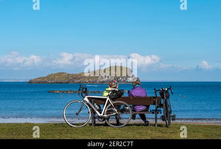 North Berwick, East Lothian, Écosse, Royaume-Uni, 28 février 2021. Météo au Royaume-Uni : soleil de printemps. Beau temps sur la côte avec deux femmes cyclistes sur un banc et admirant la vue sur le front de mer de West Bay, surplombant la Firth of Forth et l'île Craigleith Banque D'Images