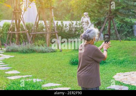 Vue arrière de la femme âgée portant un masque chirurgical pour prévenir Virus Corona (Covid-19) et photoshoot par téléphone mobile en étant debout jardin.concept de Banque D'Images