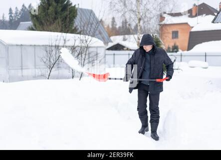 Homme adulte vêque de vêtements d'hiver noirs, qui pele de la neige dans le jardin depuis un trottoir ou une allée avec une pelle à neige rouge légère, par temps froid et ensoleillé. Banque D'Images