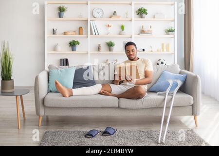 Homme noir millénaire avec jambe en plâtré assis sur un canapé, livre de lecture à la maison, paire de béquilles à proximité Banque D'Images