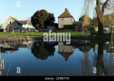 East Dean village étang avec des reflets dans l'eau des arbres et des chalets lors d'une journée d'hiver ensoleillée et calme. Banque D'Images