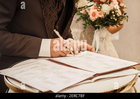 La main du marié dans un costume marron tient un stylo et met une signature sur le consentement du mariage de près. Banque D'Images