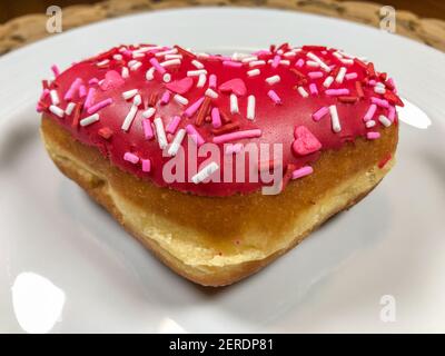 Un donut en forme de coeur rempli de pâte à gâteau, trempé dans le glaçage rouge, et recouvert de saupoudrés de Saint-Valentin festifs Banque D'Images