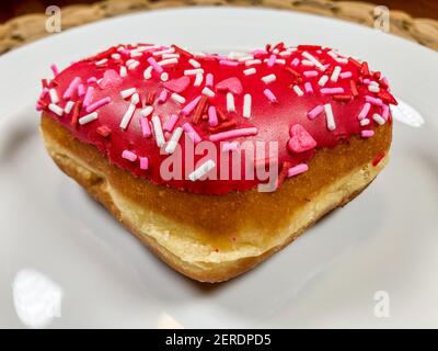 Un donut en forme de coeur rempli de pâte à gâteau, trempé dans le glaçage rouge, et recouvert de saupoudrés de Saint-Valentin festifs Banque D'Images